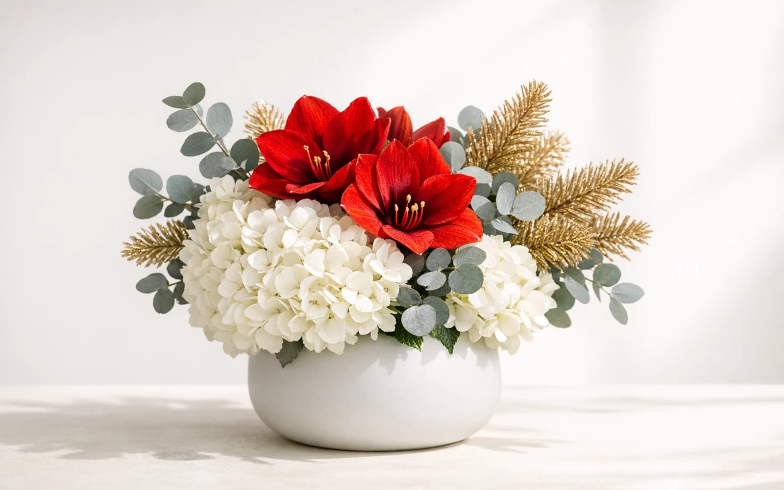 Modern holiday centerpiece with white hydrangea, red amaryllis, silver-gray eucalyptus, and gold-dipped pine branches in a matte ceramic vessel
