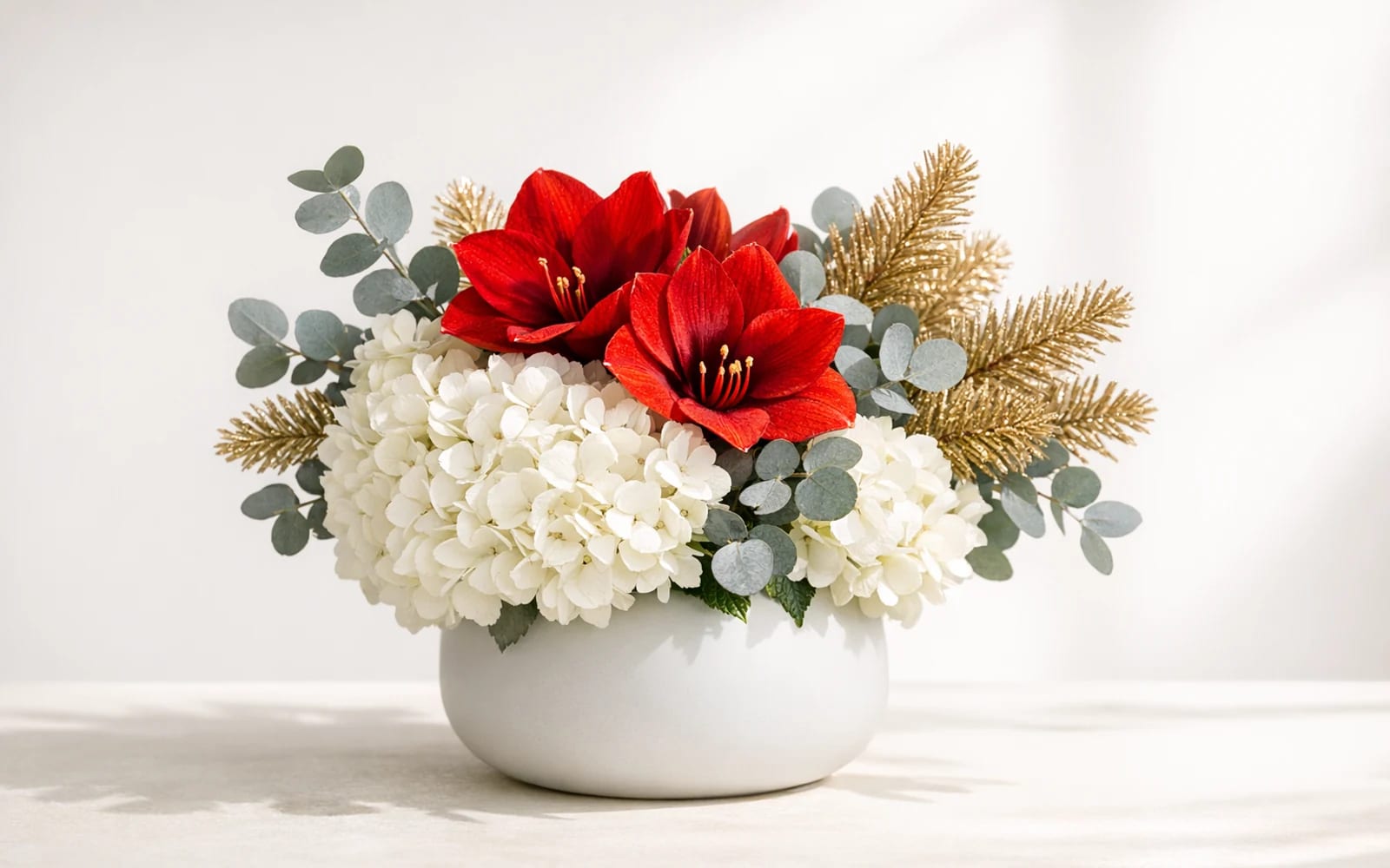 Modern holiday centerpiece with white hydrangea, red amaryllis, silver-gray eucalyptus, and gold-dipped pine branches in a matte ceramic vessel