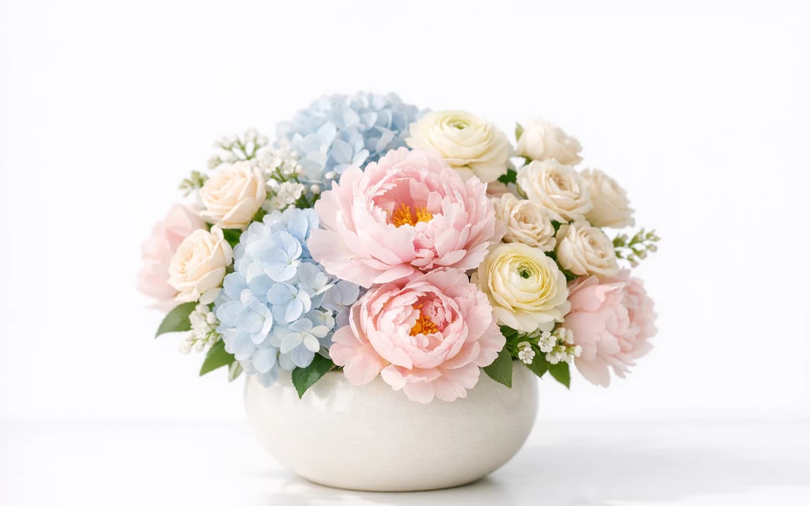 Soft pastel arrangement with blush pink peonies, powder blue hydrangea, cream spray roses, and pale yellow ranunculus in a ceramic bowl
