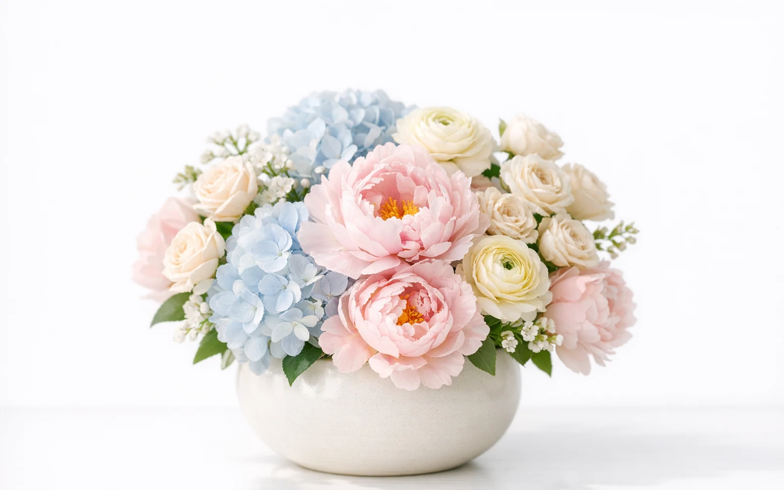Soft pastel arrangement with blush pink peonies, powder blue hydrangea, cream spray roses, and pale yellow ranunculus in a ceramic bowl