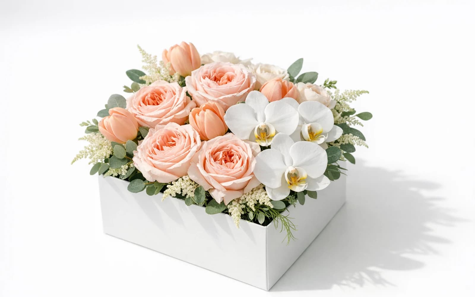 Luxurious modern square flower box with blush garden roses, peach tulips, white orchids, and textural greenery on white backdrop
