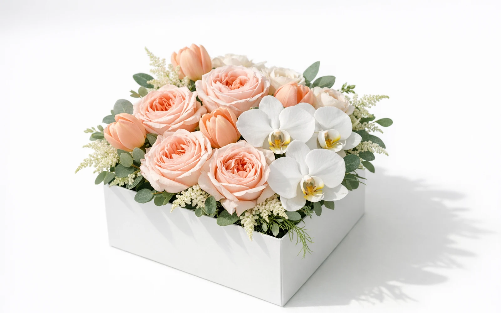 Luxurious modern square flower box with blush garden roses, peach tulips, white orchids, and textural greenery on white backdrop