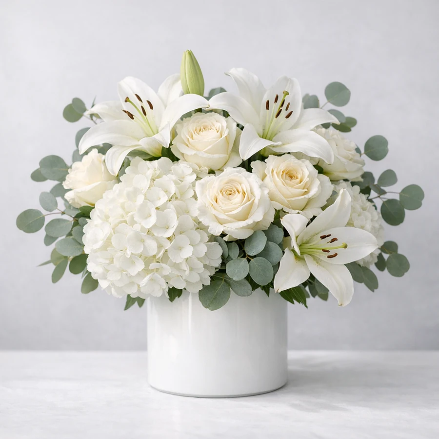 Dignified all-white sympathy floral arrangement with lilies, ivory garden roses, hydrangea, and eucalyptus in a clean white ceramic vessel
