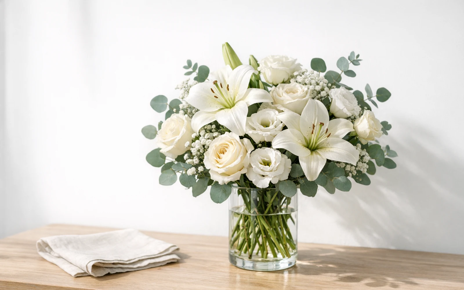Dignified all-white sympathy floral scene with lilies, garden roses, and eucalyptus on a simple wooden table, styled with quiet reverence by LiNa Garden